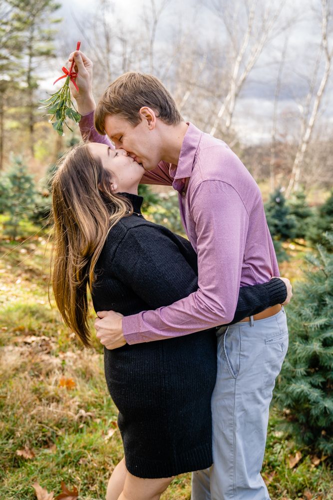 A romantic couple kisses under mistletoe in a Christmas tree farm during a winter engagement photoshoot.