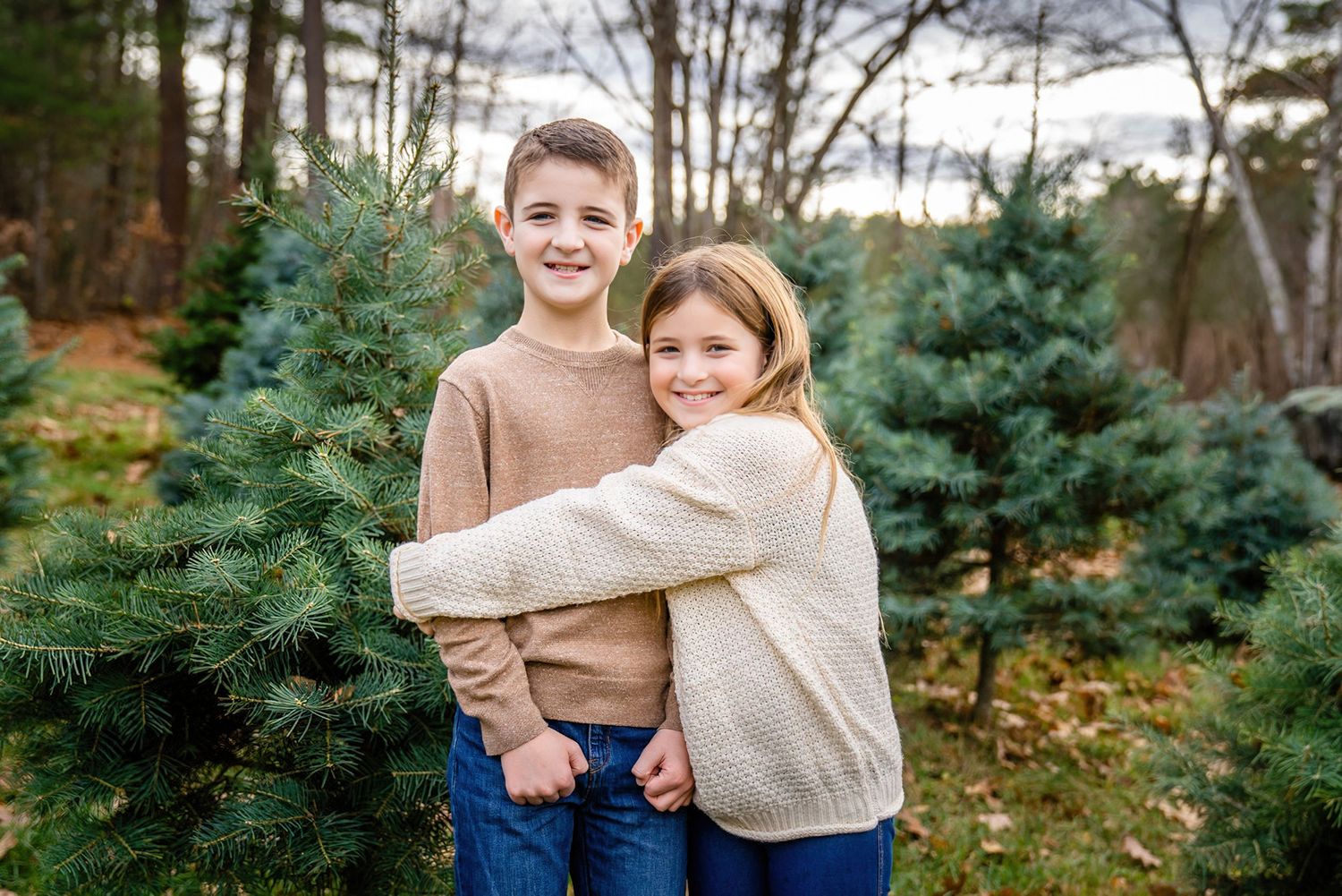 Two children hug and smile while standing among evergreen Christmas trees at a tree farm on an autumn day.