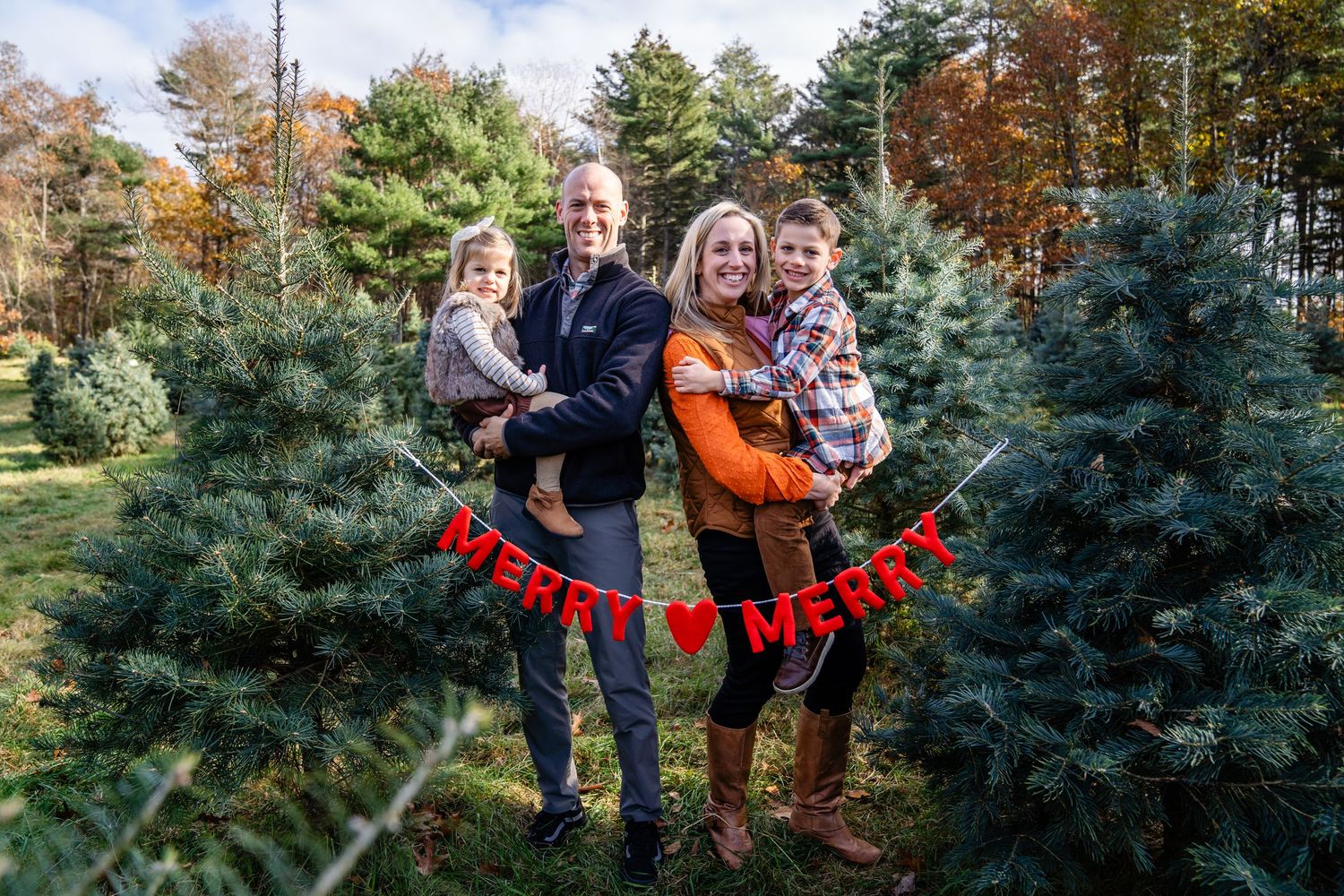 A festive holiday portrait among evergreen Christmas trees with a red "Merry Christmas" banner display.