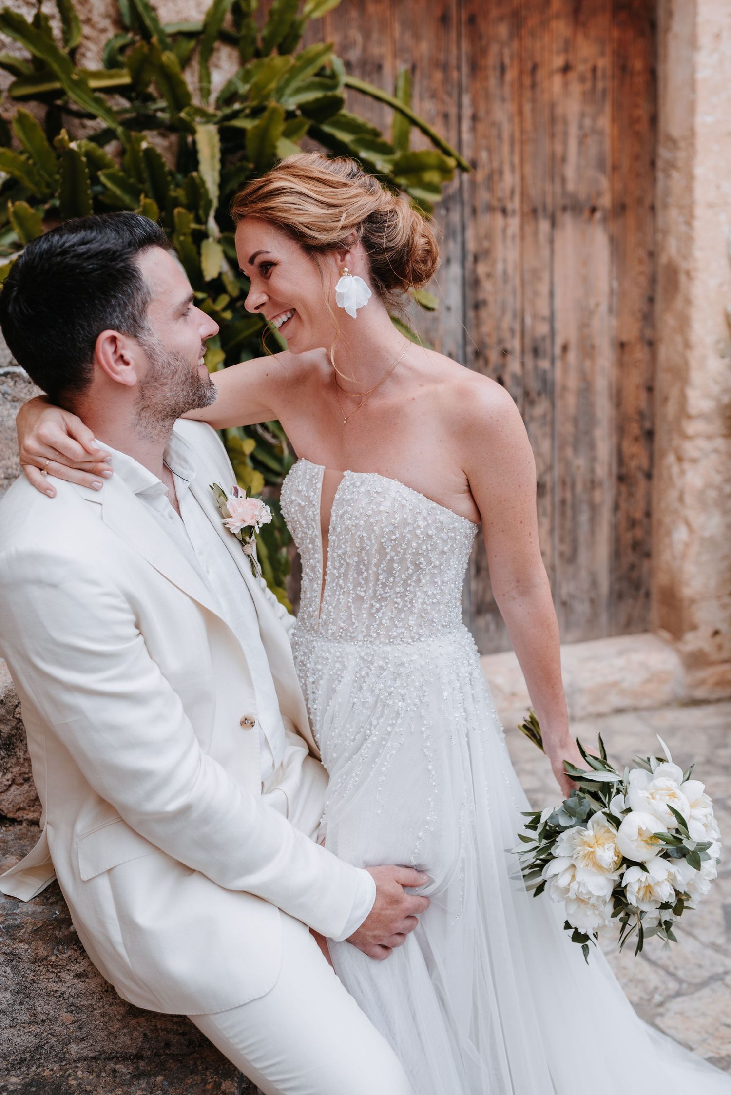 Ein romantisches Hochzeitspaar in weißer Kleidung umarmt sich und genießt intime Momente vor mallorquinischen Holztüren in der Finca Xiclat.