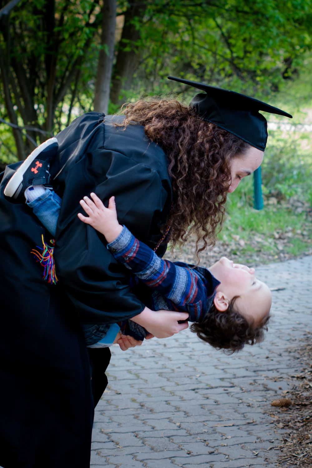A graduate in cap and gown shares a joyful moment dipping their dance partner on a tree-lined path.