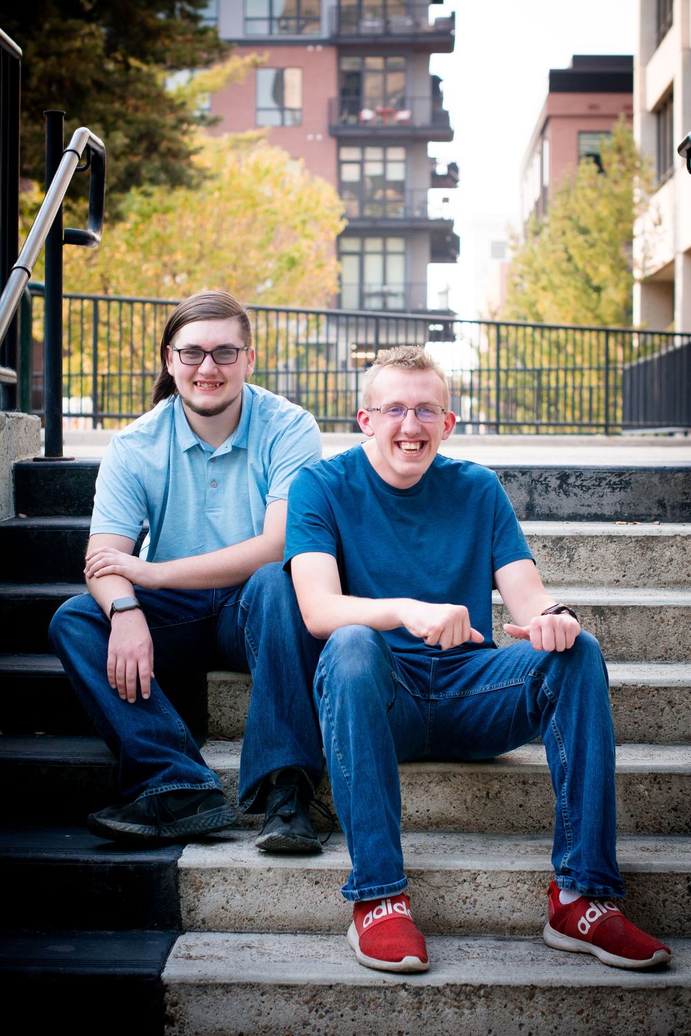 Two people in casual attire sitting on concrete steps in an urban setting with autumn trees and buildings in background.