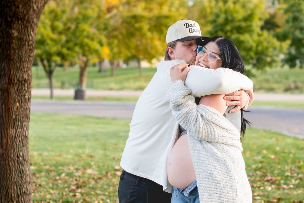 Couple embracing outdoors during maternity photo shoot with fall foliage in background.