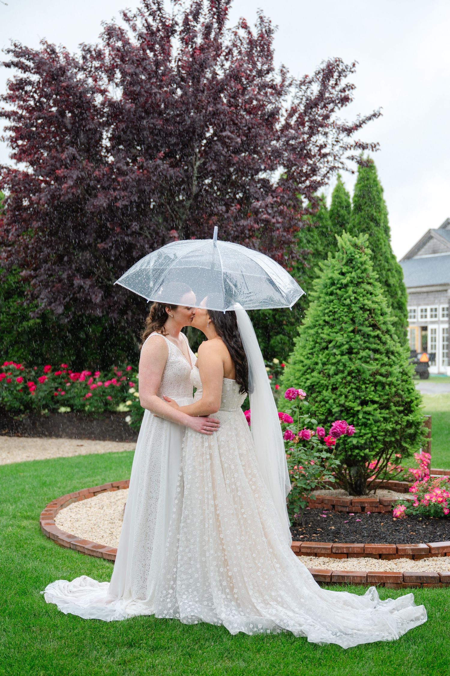 Two people in white wedding gowns hold hands on a wooden porch with a scenic green background.
