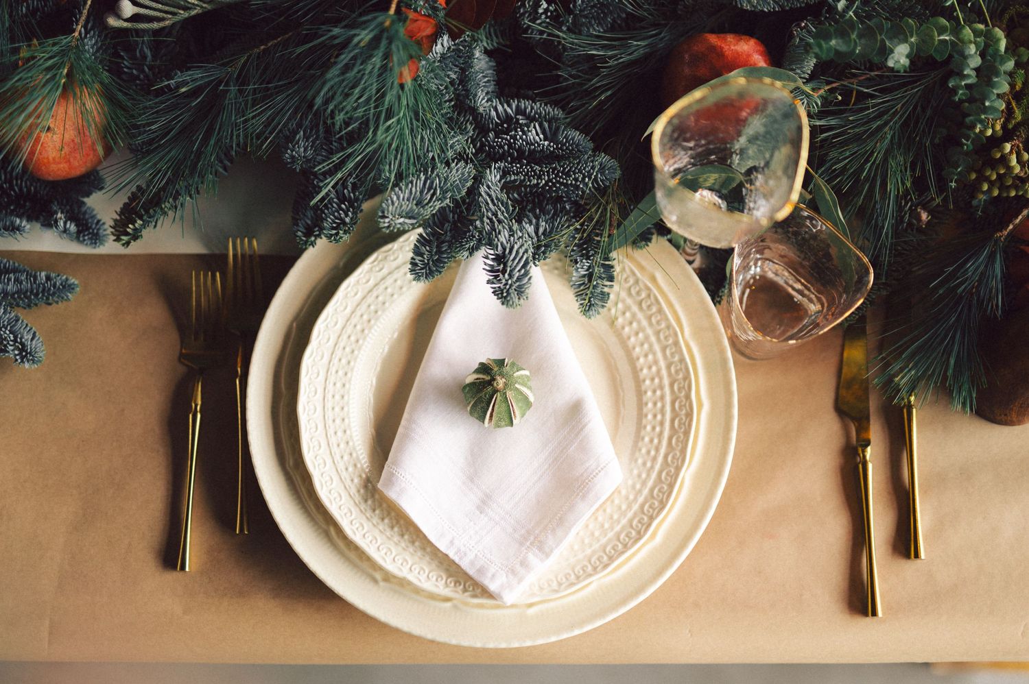 Festive holiday place setting with white plates and evergreen branches on kraft paper with gold silverware.