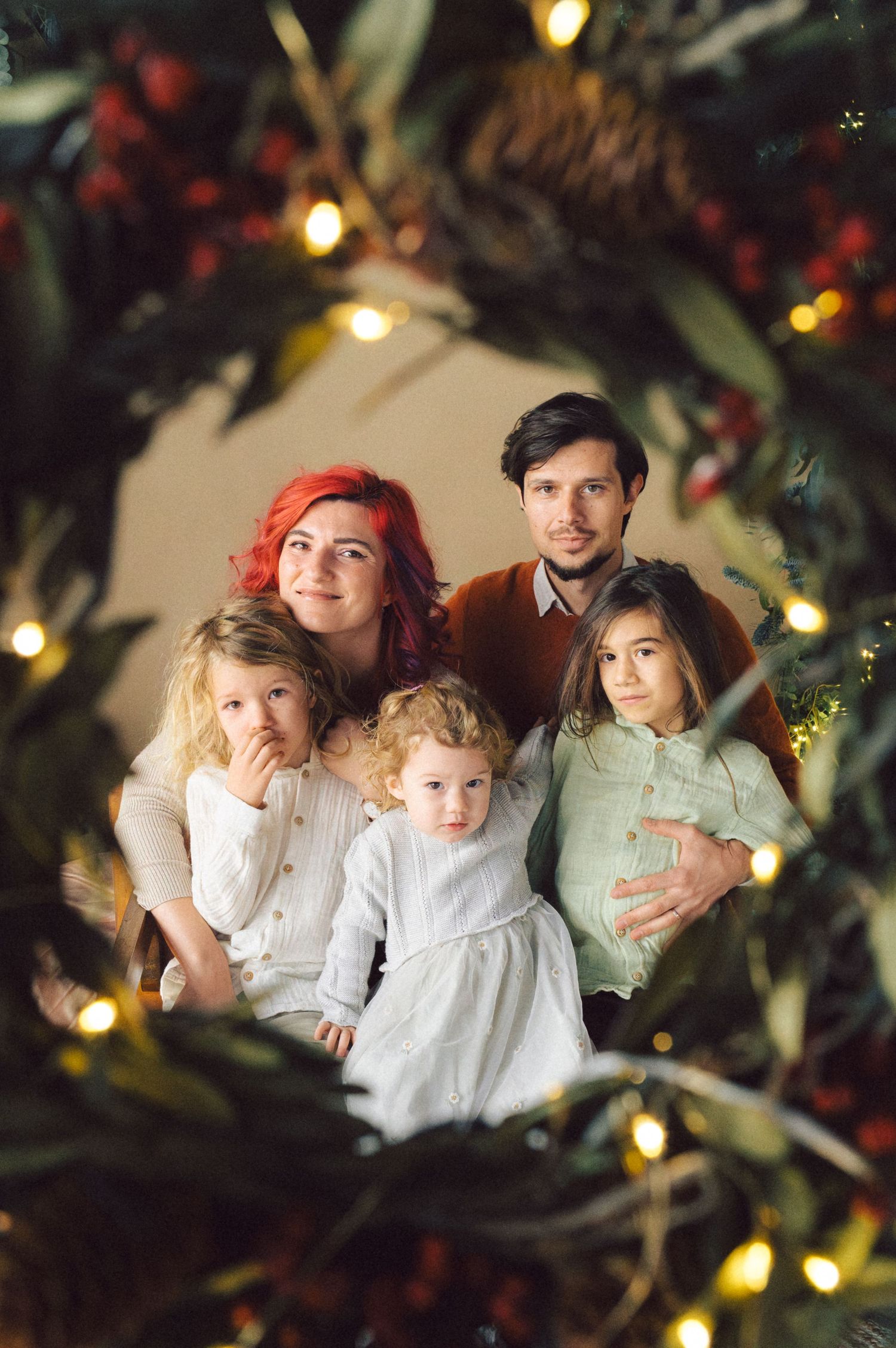 A holiday family portrait framed by evergreen branches and twinkling lights in warm ambient lighting.