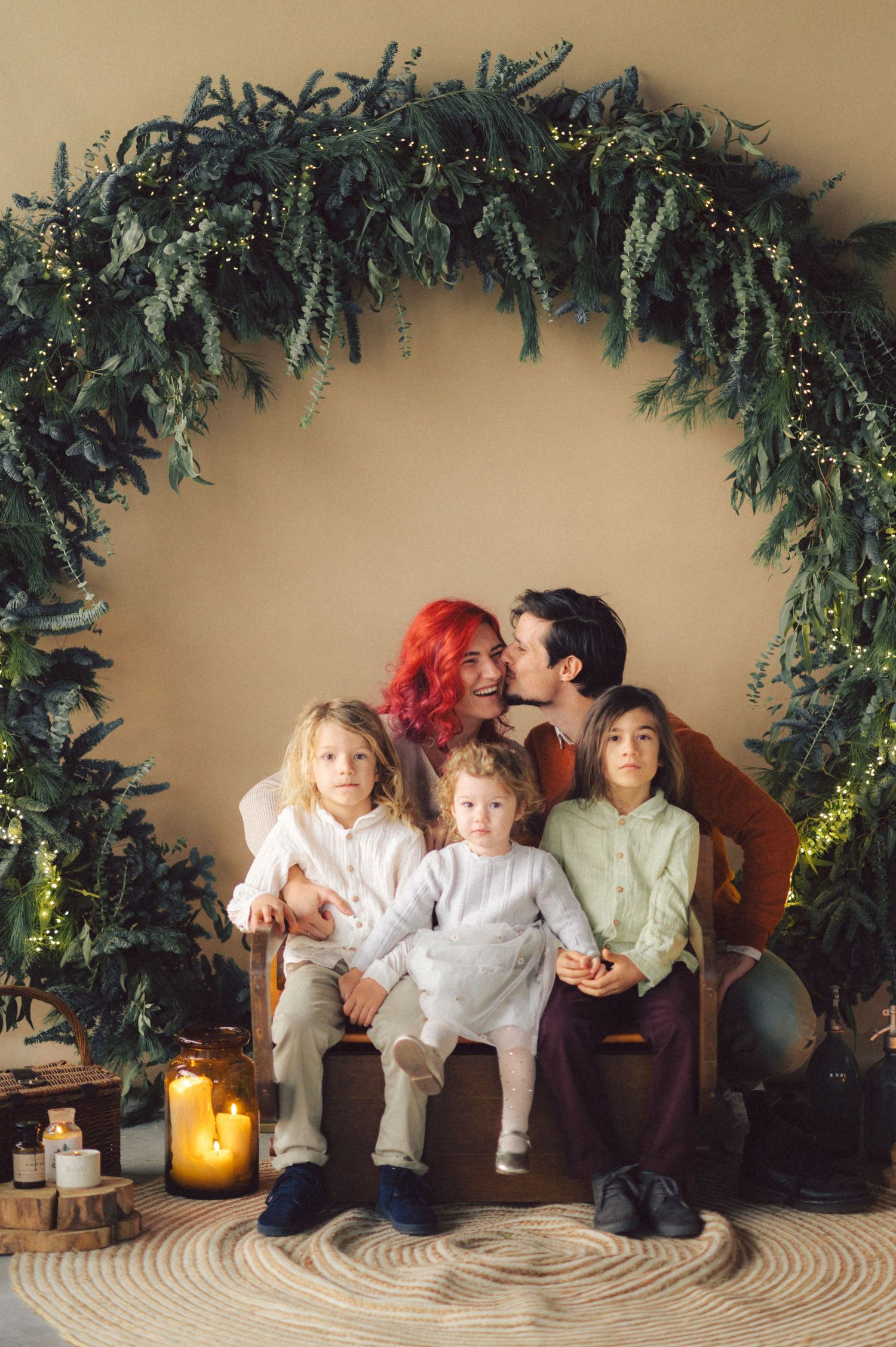A group sits together under a large botanical arch decorated with evergreen foliage and lit by glowing candles on the floor.