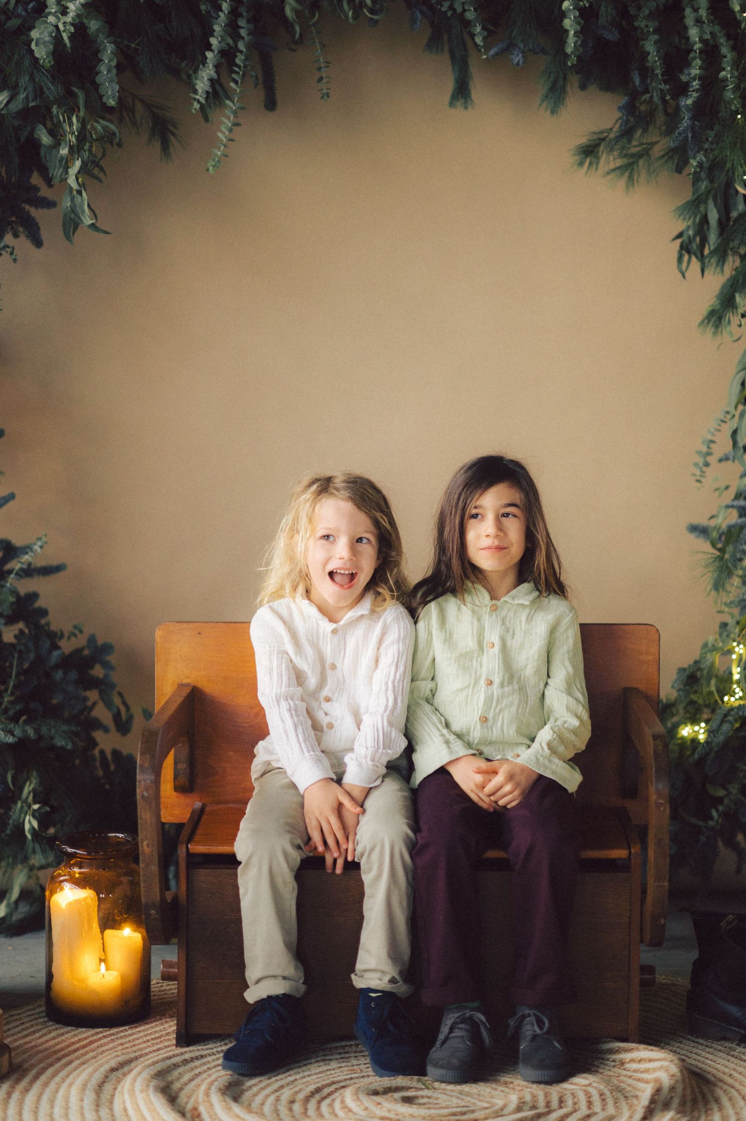 Two children sitting on a wooden bench surrounded by evergreen branches and candlelight in a cozy holiday setting.