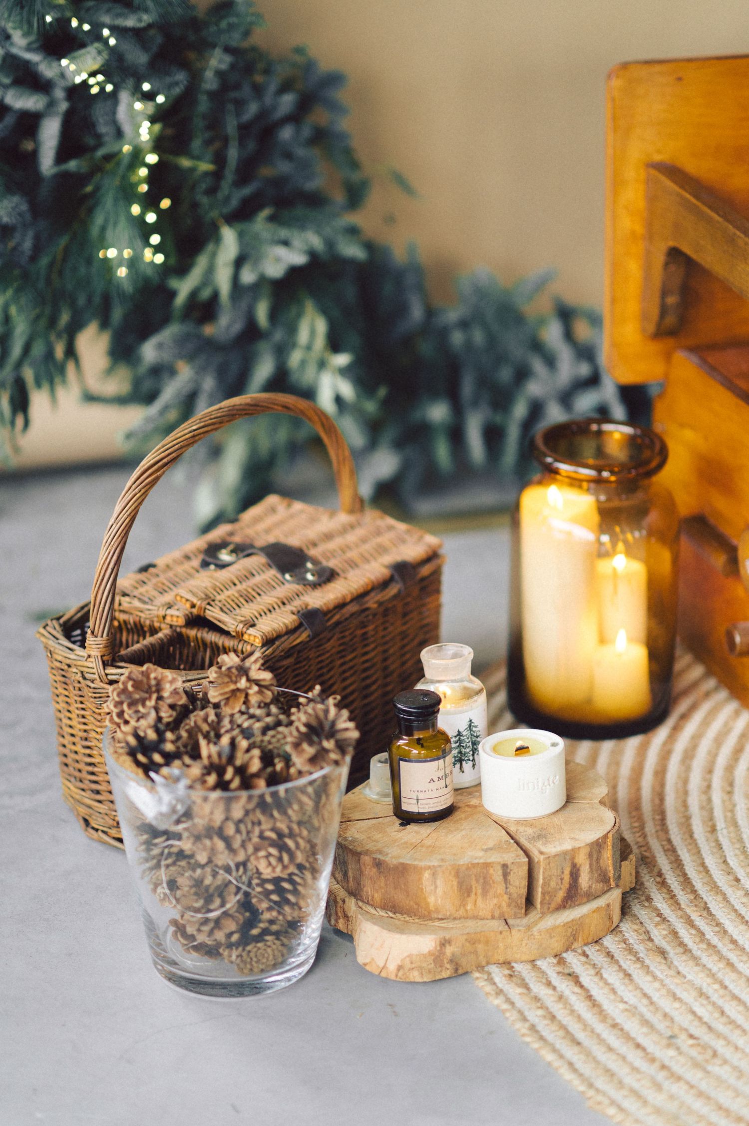 Rustic home decor display with wicker basket, pinecones, wood slices, glass jars and candlelight against greenery backdrop.