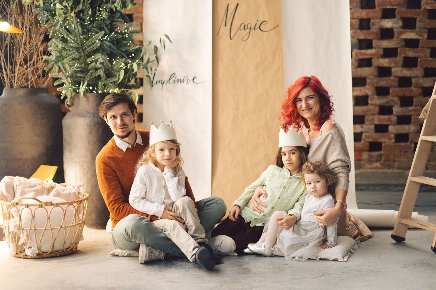 A family dressed in white and earth tones sits together on a patio with brick wall and potted plants in background.