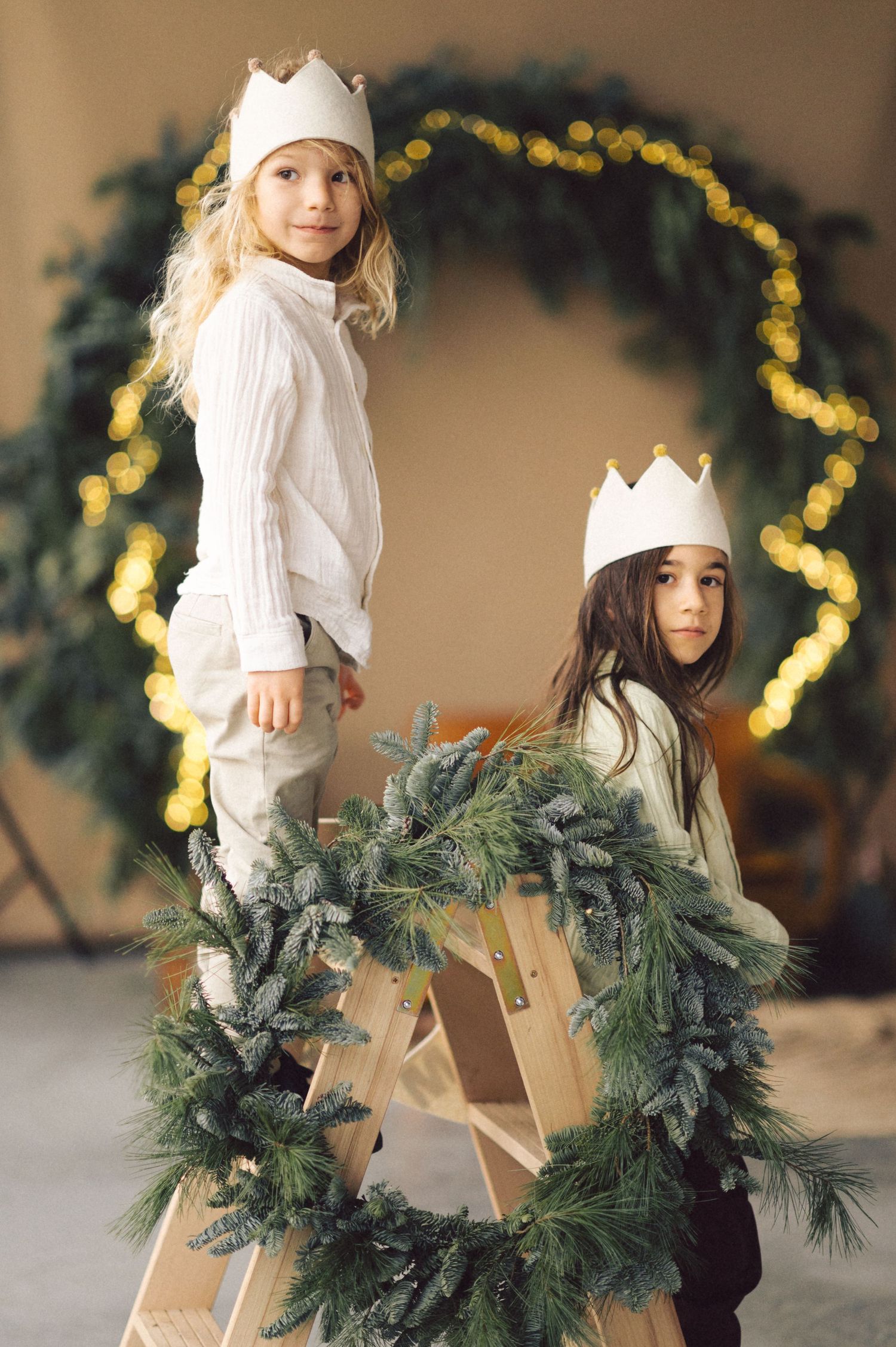 Two children wearing white paper crowns stand on a ladder with an evergreen wreath and heart-shaped lights in the background.