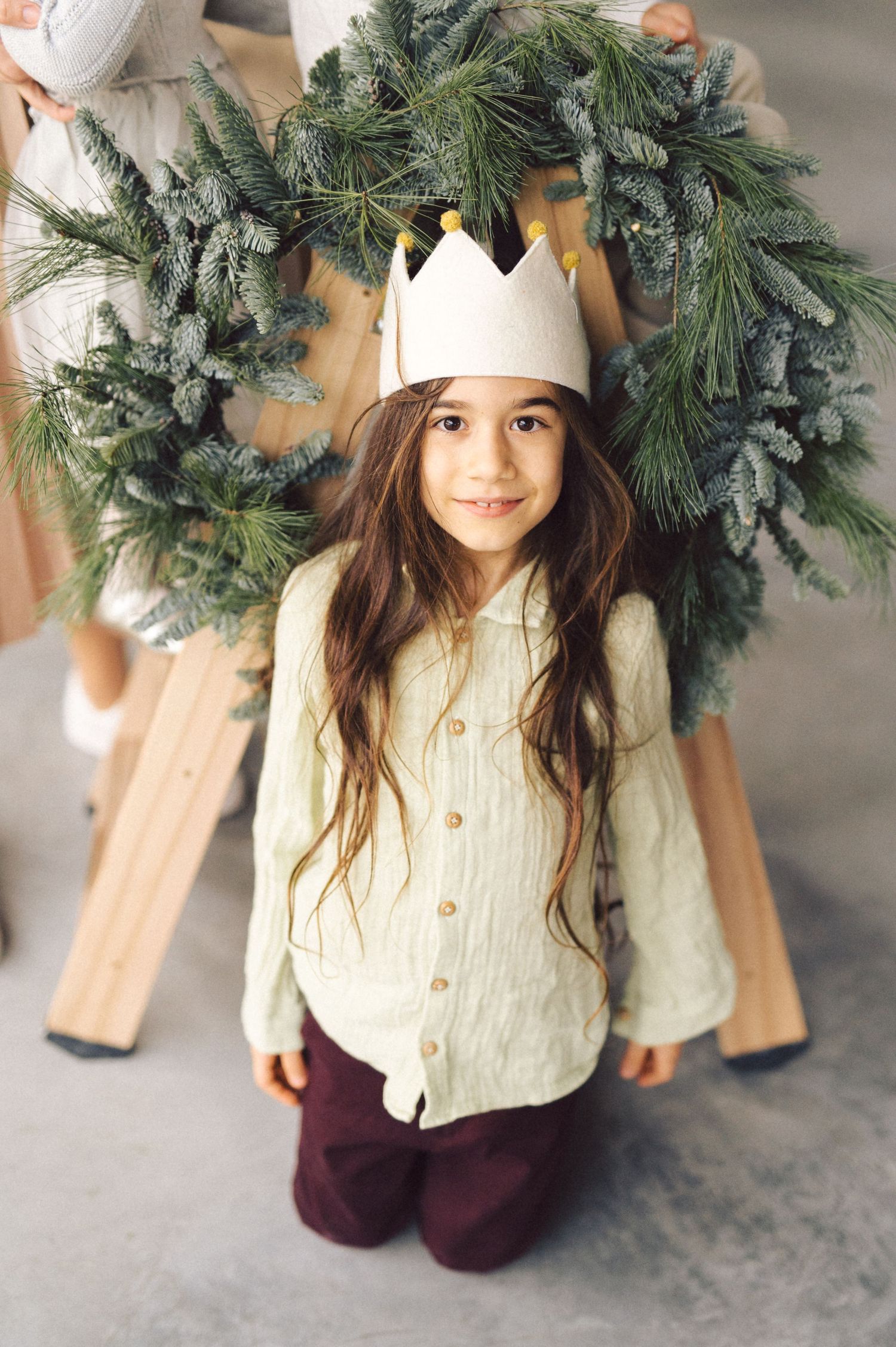 A festive holiday scene with an evergreen wreath and someone wearing a white paper crown and cream sweater.