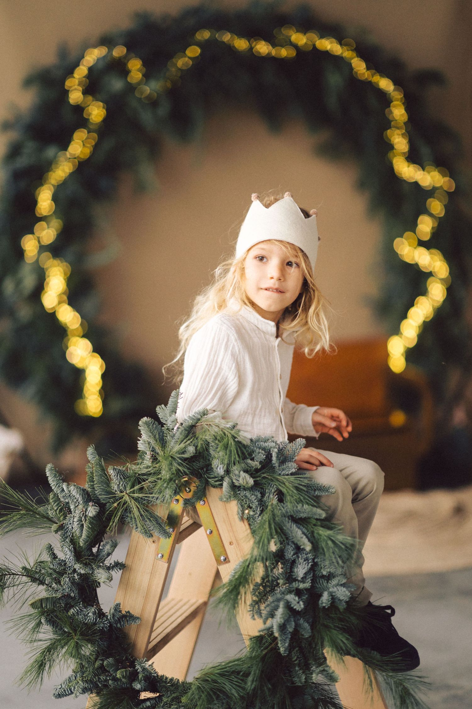 A child in white winter attire sits among holiday decorations with a lighted wreath and evergreen garland.