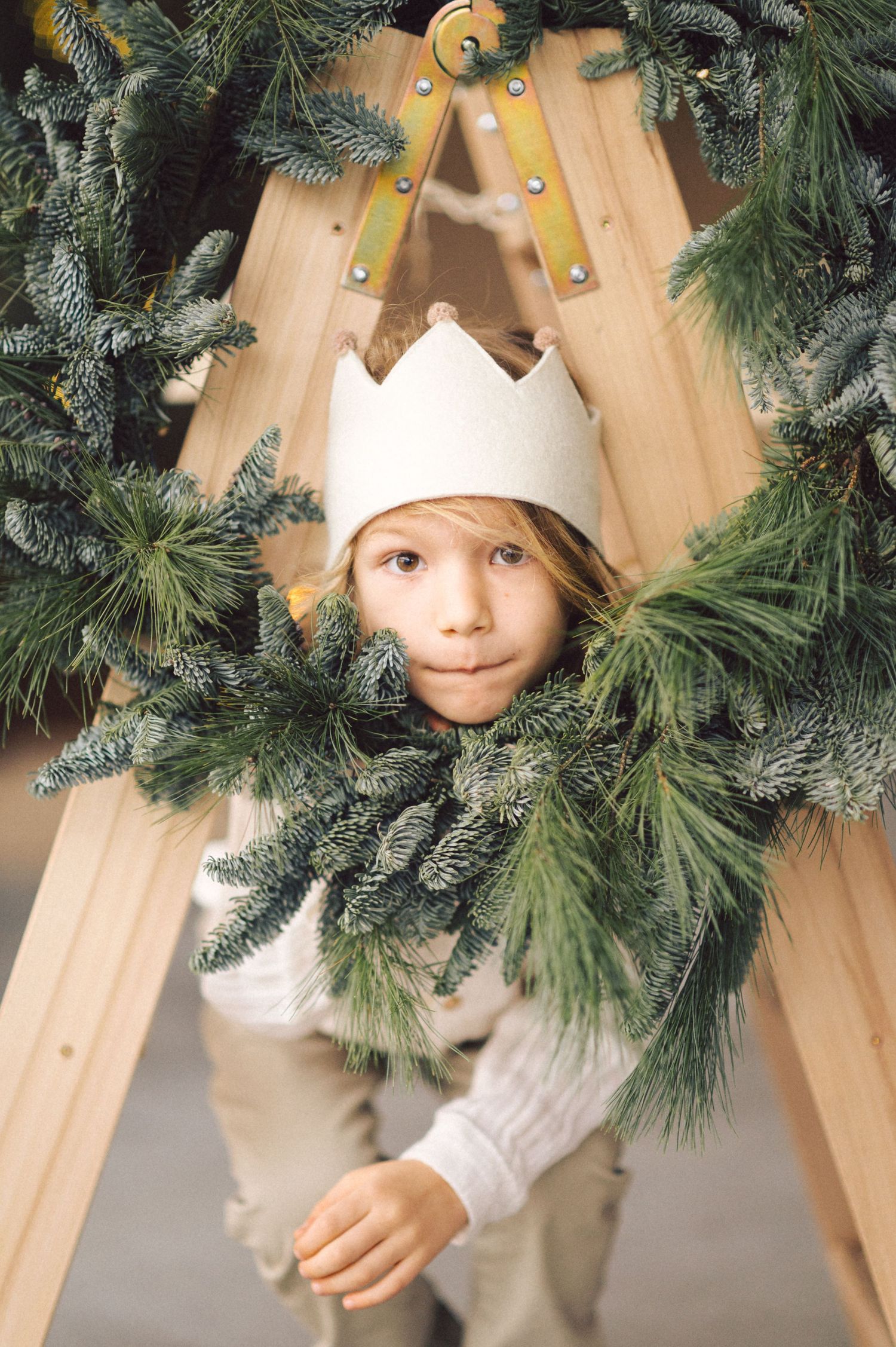 A child wearing a white paper crown peeks through evergreen branches against a wooden teepee backdrop during holiday decorating.