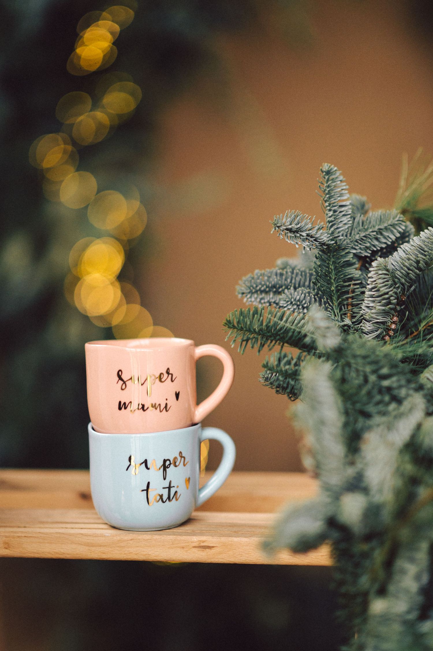White ceramic coffee mug with handwritten text sits beside evergreen branches with golden bokeh lights in background.
