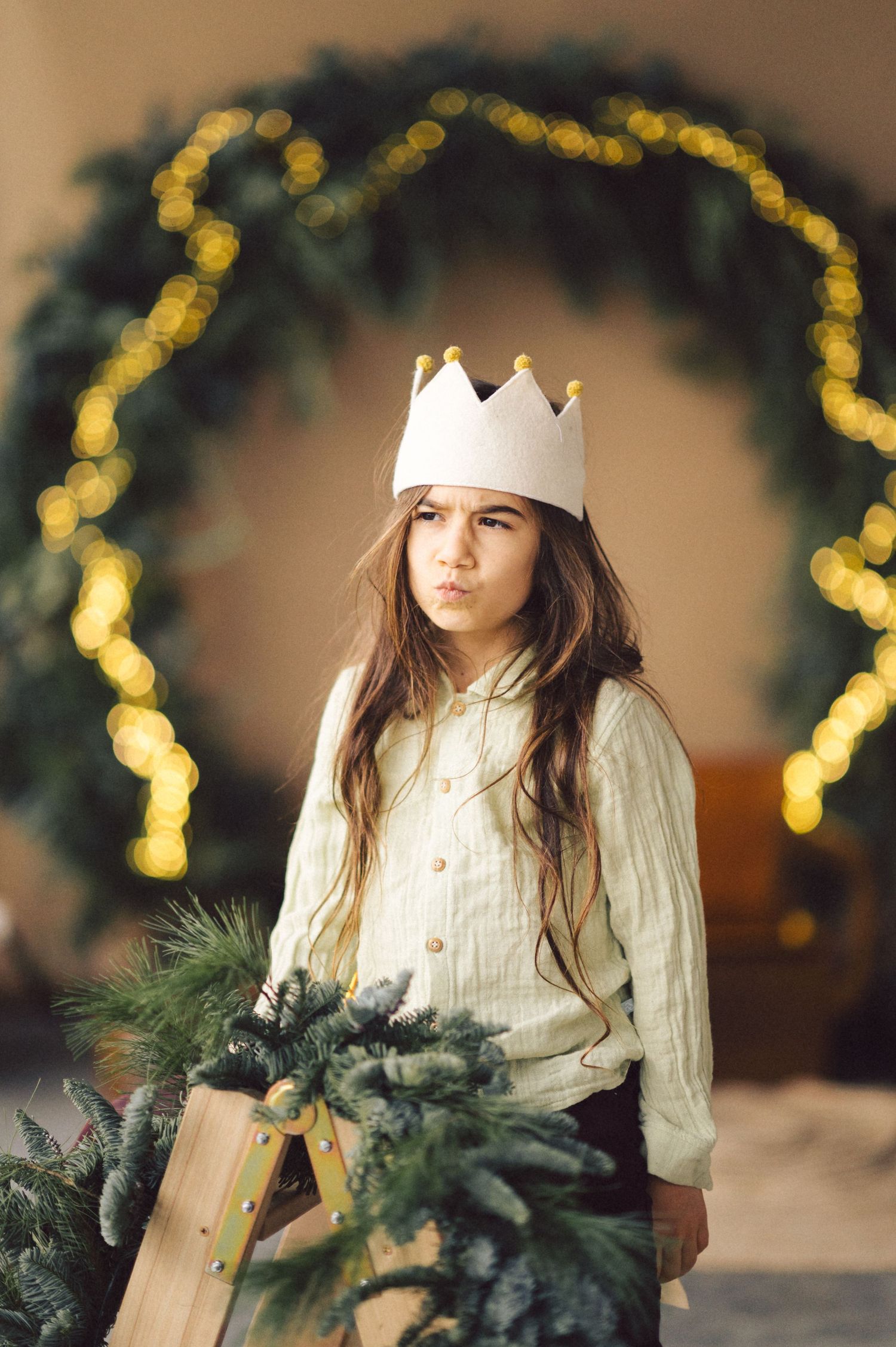 A child wearing a white knit sweater and paper crown poses with Christmas greenery in front of a glowing arch backdrop.