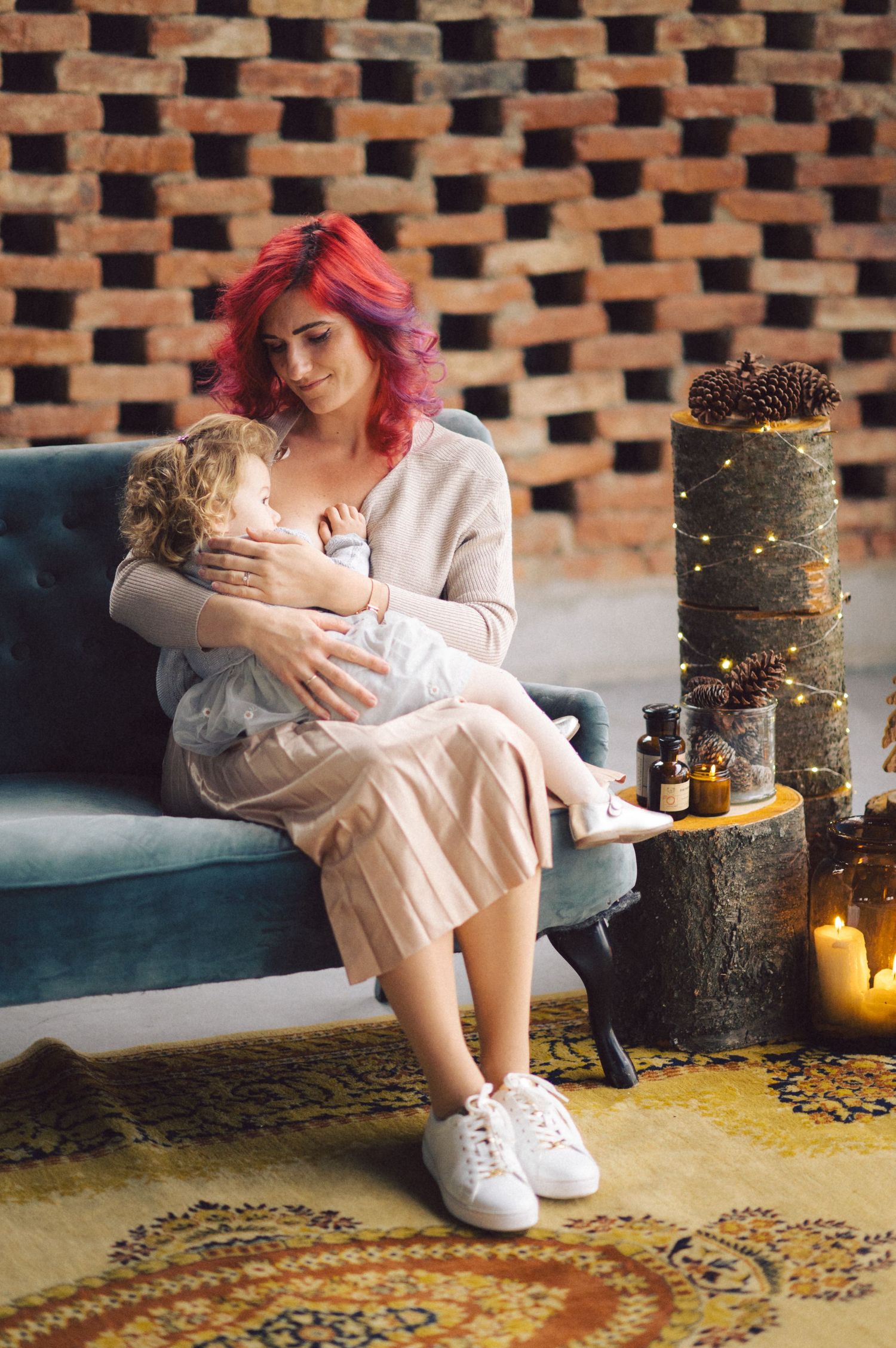 Person with pink hair in cream dress sits on blue couch with candles and decorative cake display against brick wall.