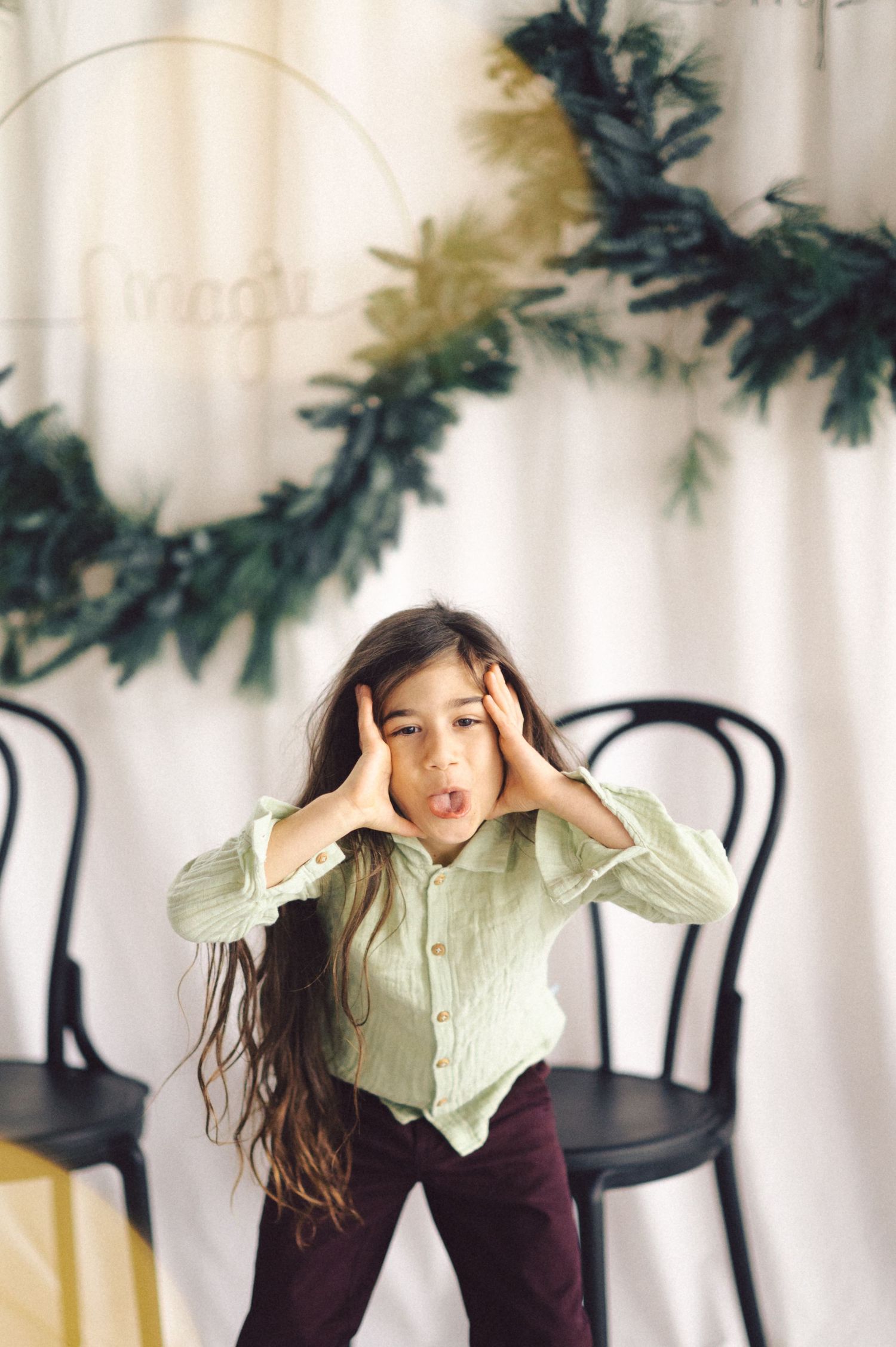Person in mint green shirt and dark pants makes expressive pose against white backdrop with holiday greenery garland.