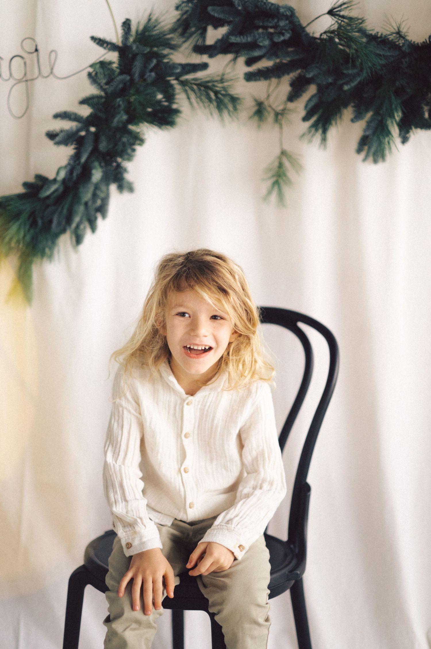 A child sits on a black chair beneath evergreen holiday garland against a white backdrop in a festive portrait setting.