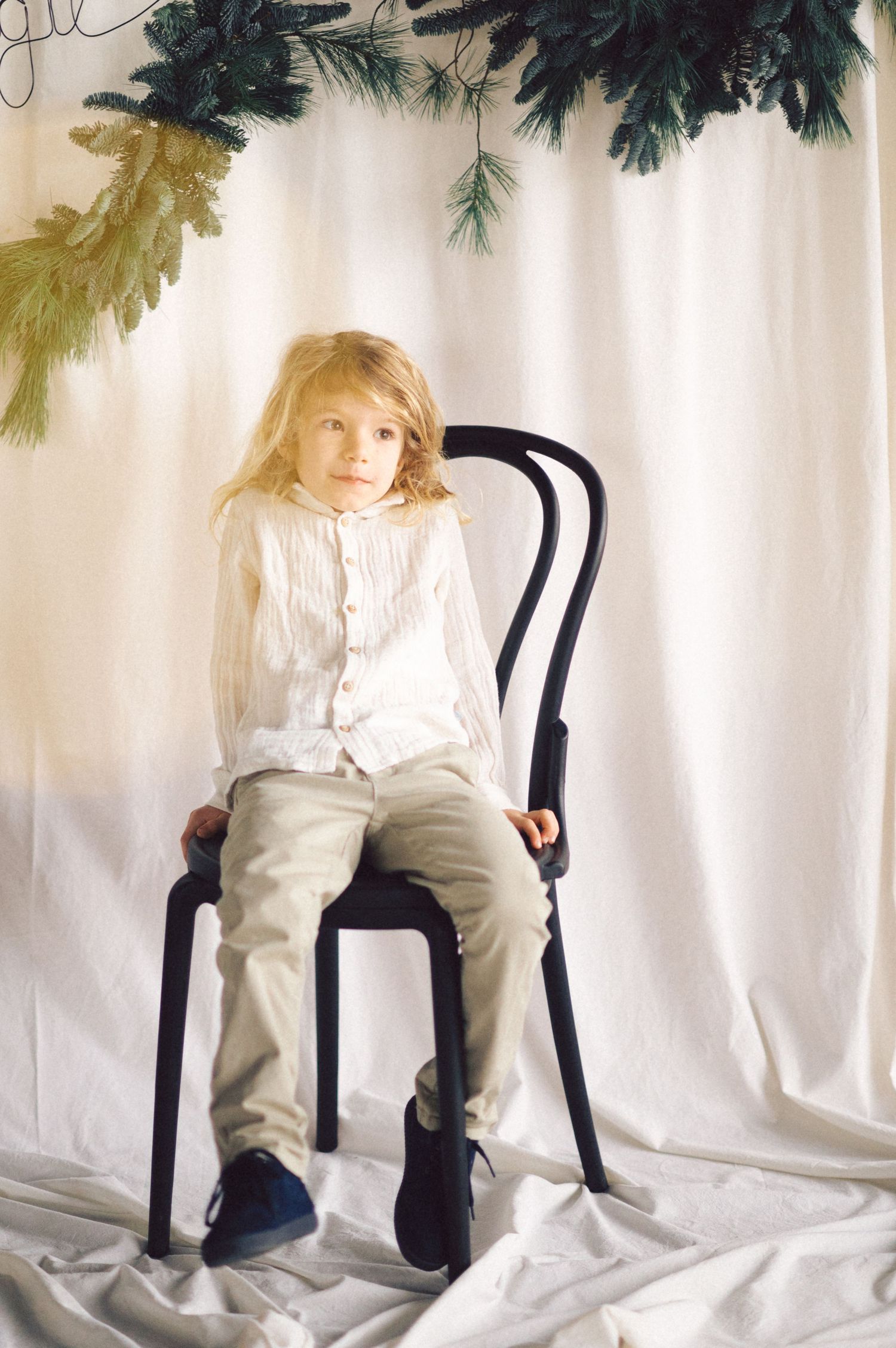 A child in a white top and khaki pants sits on a black chair against a white backdrop with hanging foliage decor.