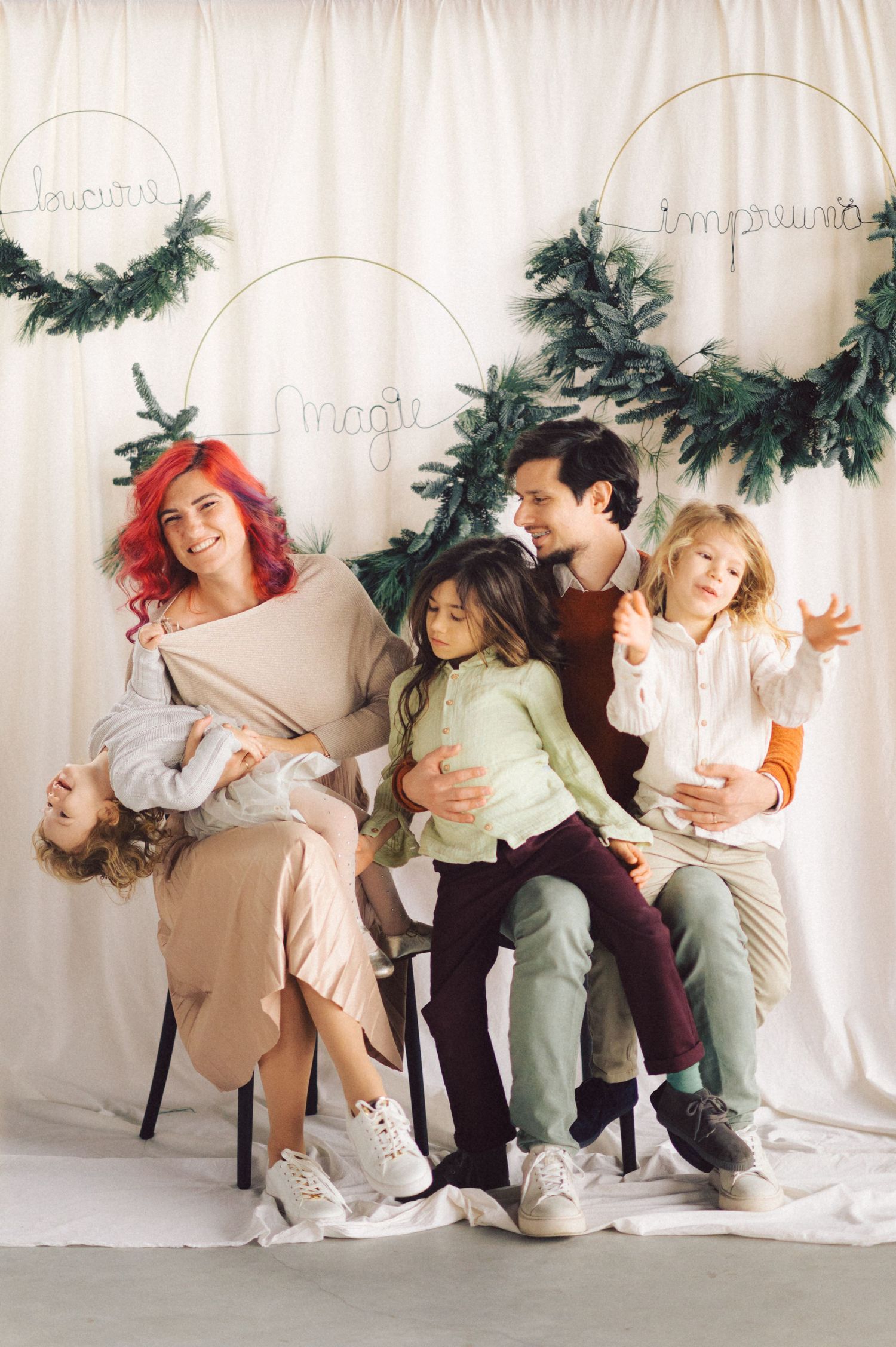 A family sits together against a white backdrop decorated with evergreen garland wreaths and holiday decor.