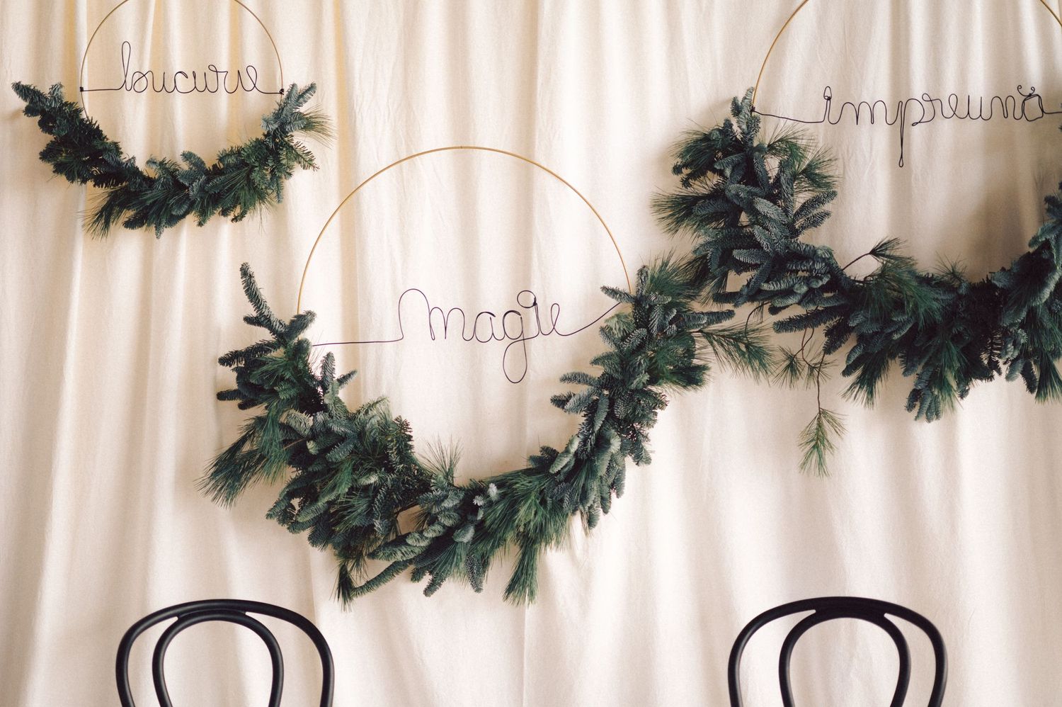 A delicate evergreen garland with light trim hangs against a white curtain backdrop above two dark wooden chairs.