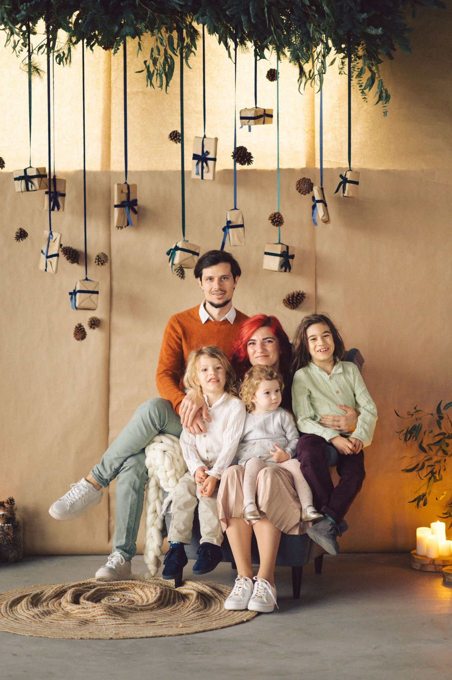 A cozy family portrait with hanging lights and candles, sitting on a round rug against a beige wall with greenery.