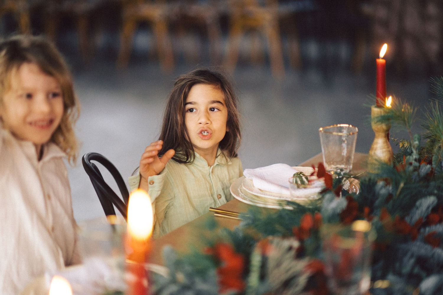 Children sit at a festive holiday dinner table with lit candles and decorative greenery.