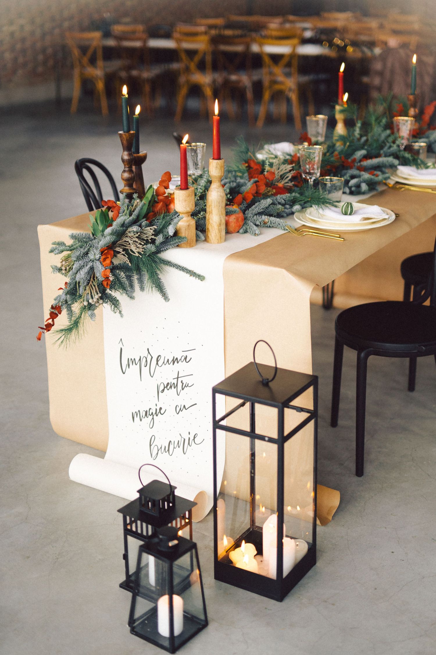 Elegant holiday table setting with kraft paper runner, evergreen garland, red candles and black lanterns on concrete floor.