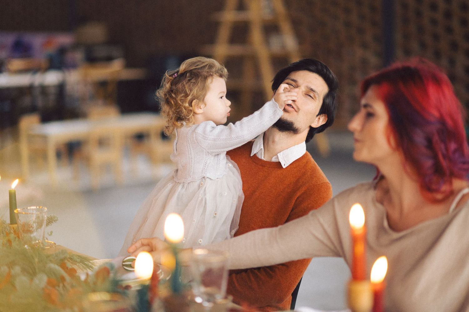 A child in a white dress reaches out while sitting at a candlelit dining table with two others during a festive dinner.
