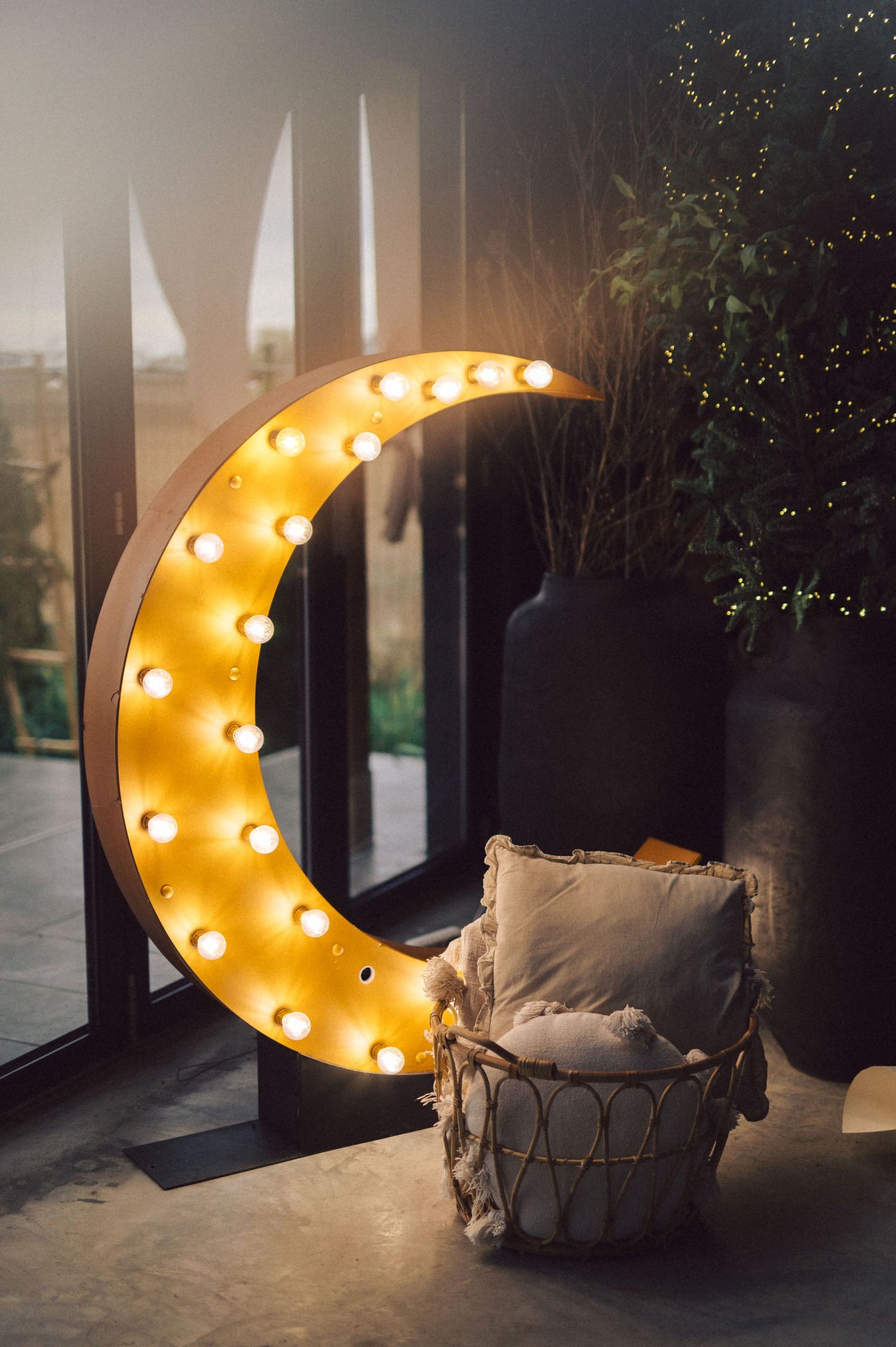 A large illuminated crescent moon light fixture glows warmly next to a cozy wicker chair with beige cushions.