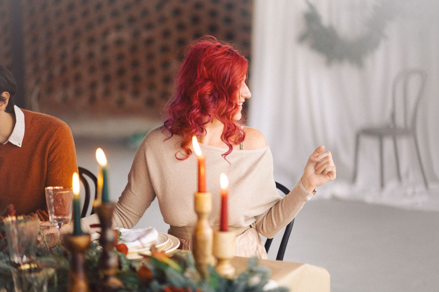 Someone with vibrant red hair sits at an elegantly set dining table with gold candlesticks and decorative elements.