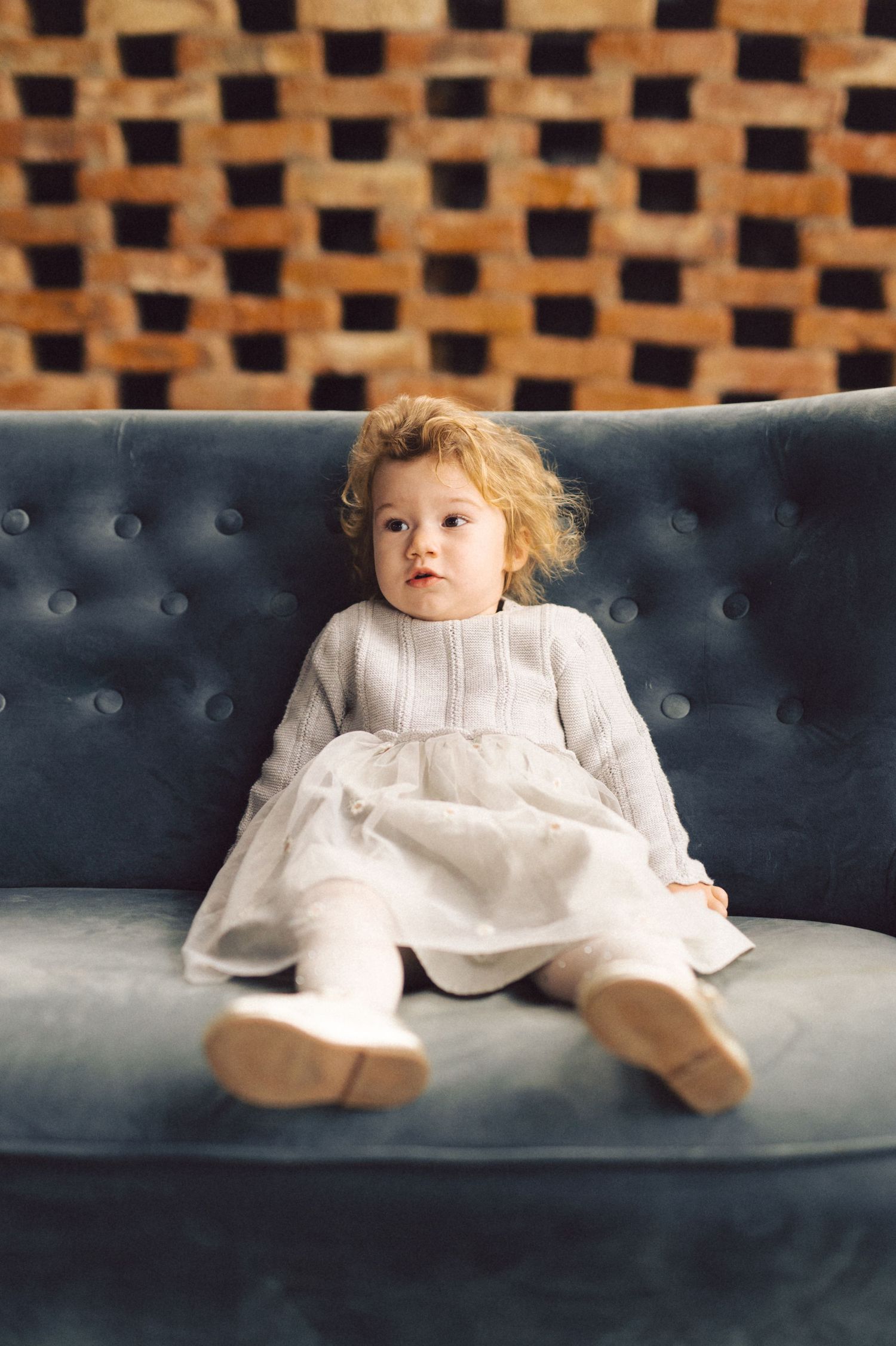 A small child wearing a white dress sits on a blue tufted sofa against a geometric patterned wall.