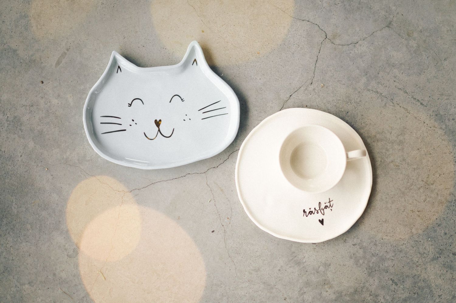 Two ceramic plates on gray surface, one shaped like a smiling cat face and one white circular plate with small heart detail.