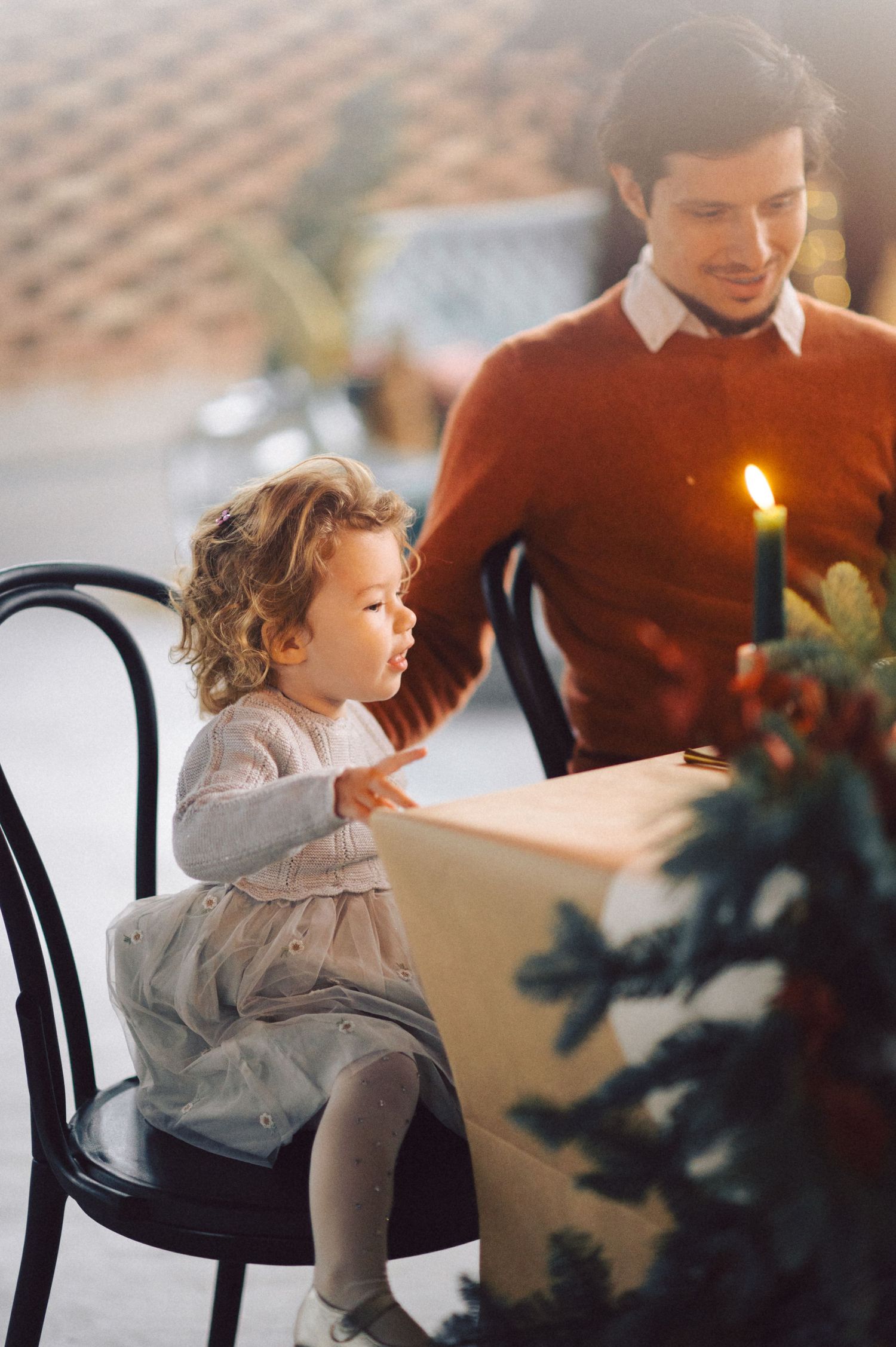 A festive holiday scene with a small child in a dress sitting near a lit candle and evergreen decorations.