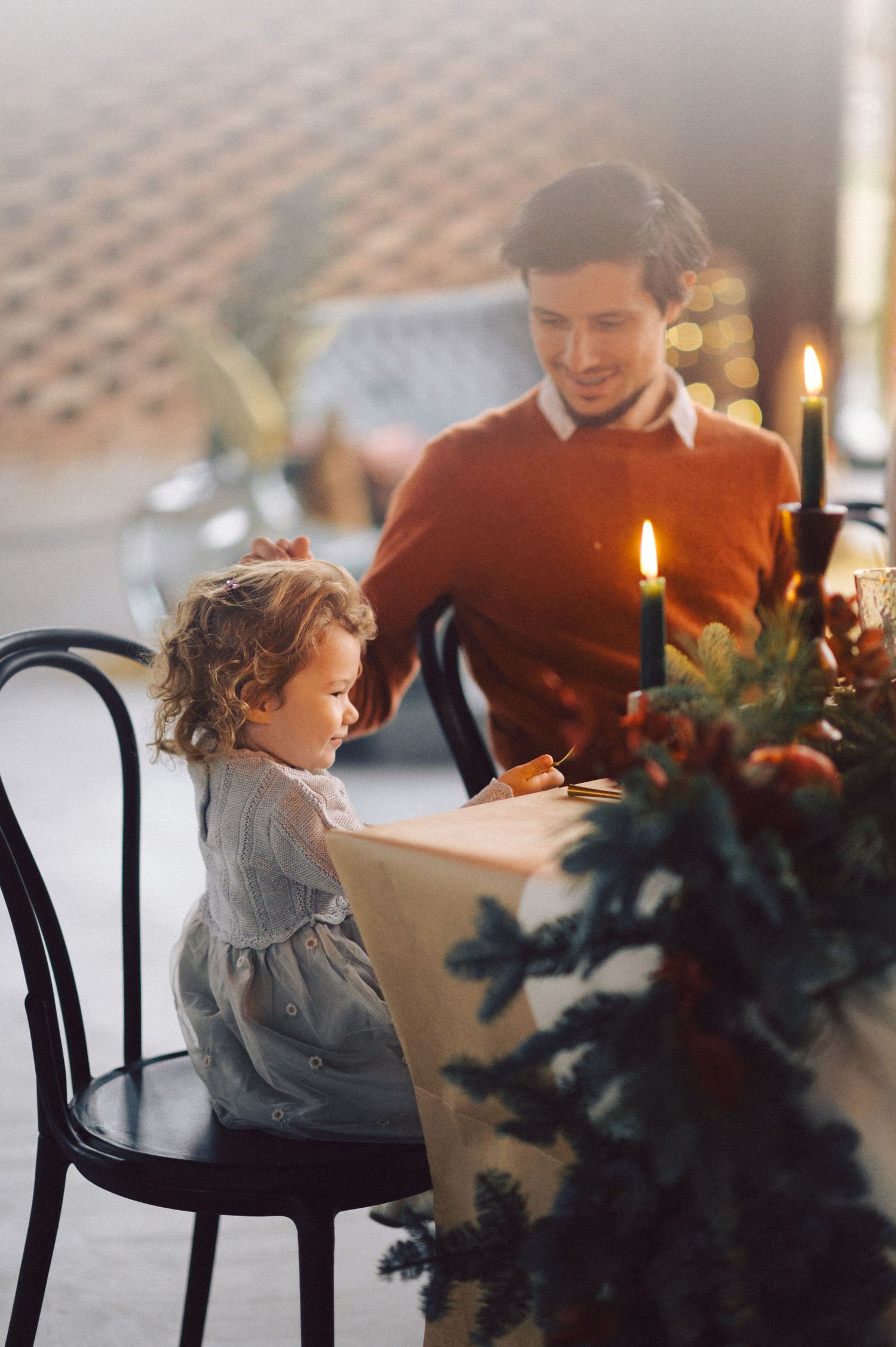 A small child in a gray dress sits at a candlelit table during a cozy holiday dinner gathering.