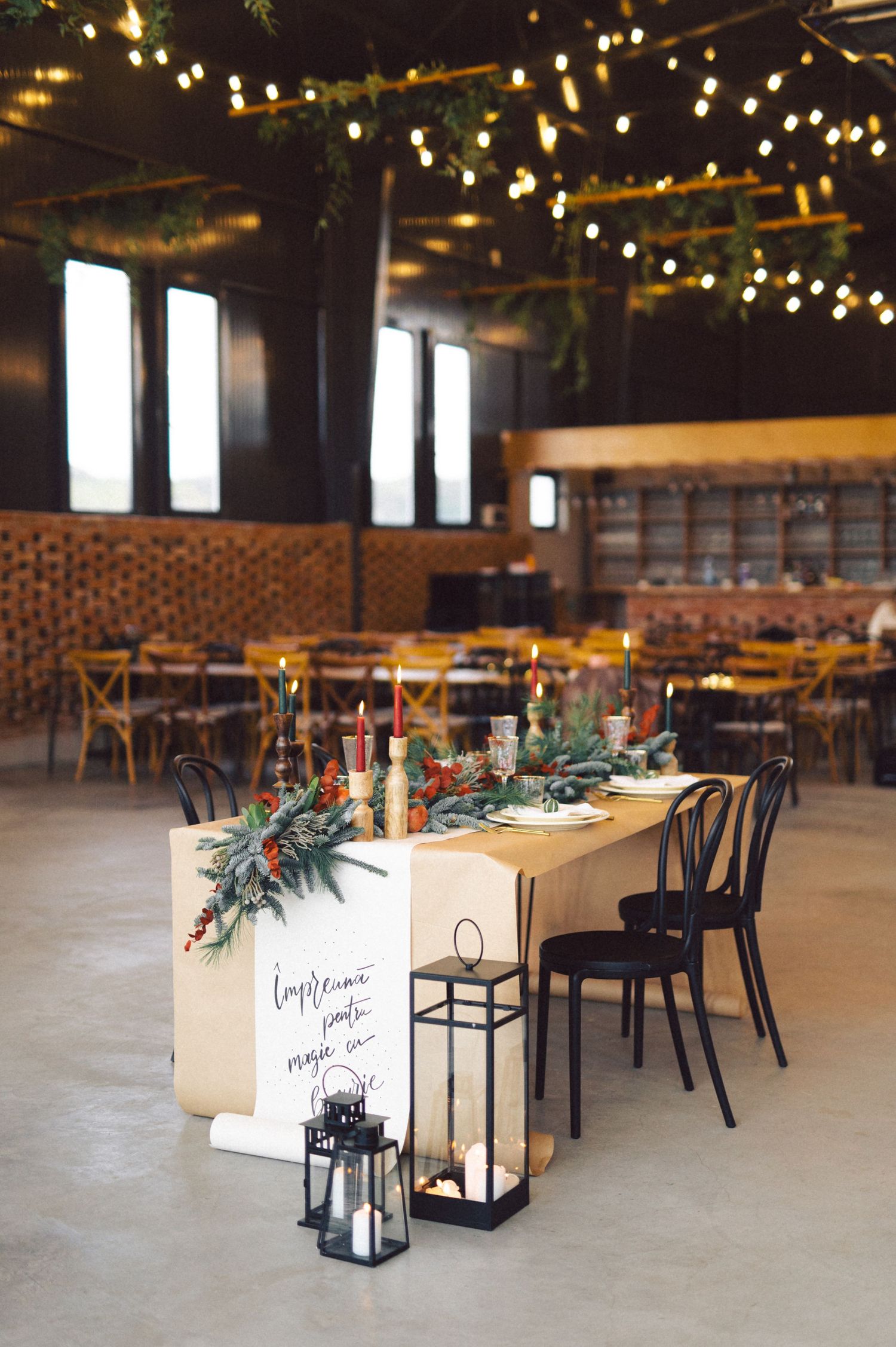Rustic indoor dining space with string lights, wooden tables, black chairs and lantern decorations.