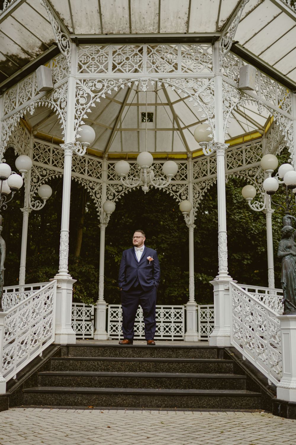 A person in a navy suit stands in an ornate white Victorian gazebo.