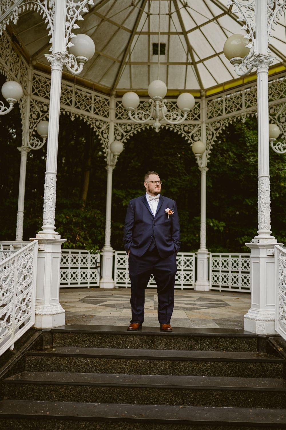 A person poses in formal attire inside a decorative white gazebo with globe lights.