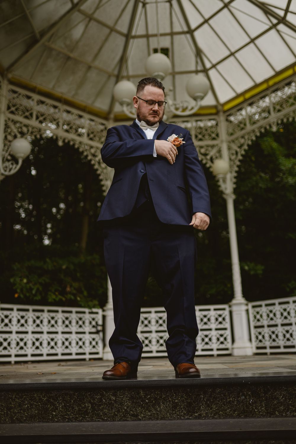 Wedding guest in navy suit checks watch while standing in ornate white gazebo at outdoor venue.