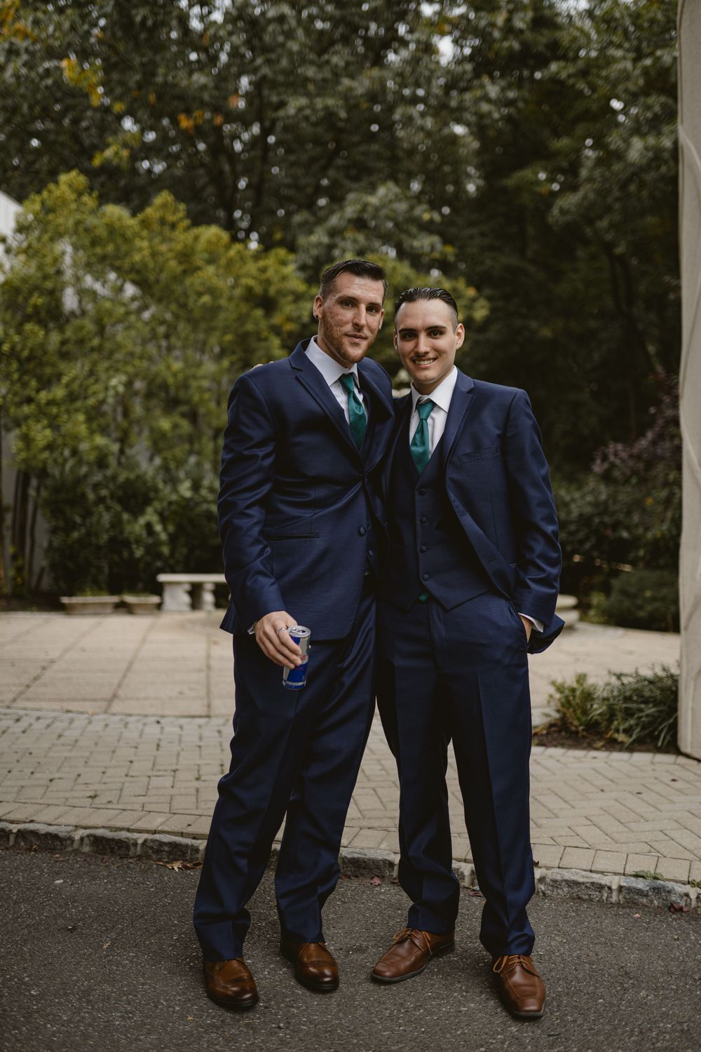 Two people in matching navy suits and teal ties pose together at outdoor wedding venue.