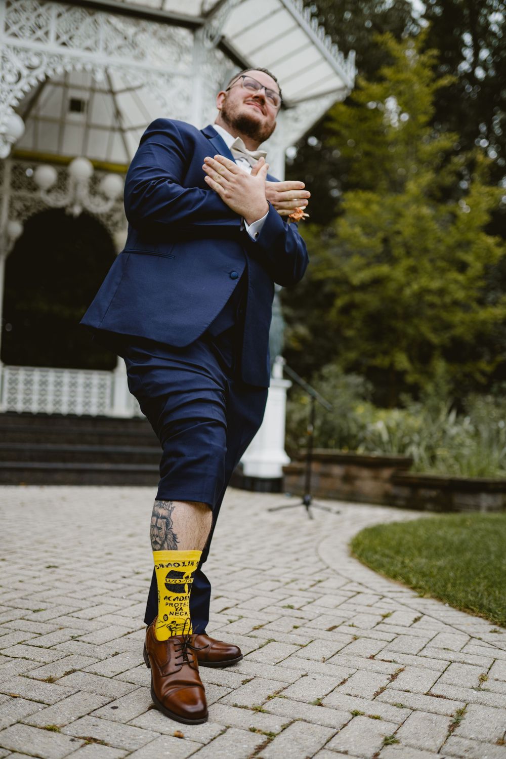 Person in navy suit expressing joy with hand on chest at outdoor wedding venue.