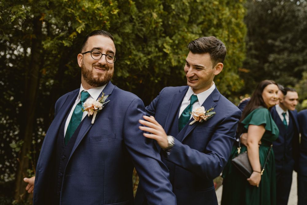 Two groomsmen share a candid moment at an outdoor wedding celebration wearing navy suits and green ties.