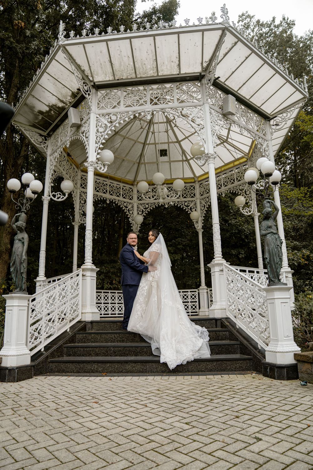 A couple poses for photos under an ornate white Victorian style wedding gazebo with globe lights.