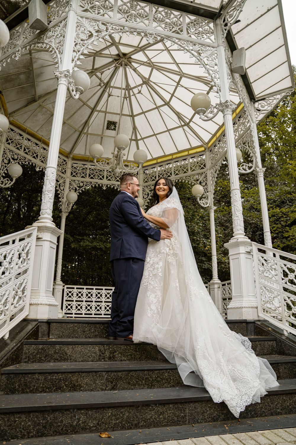 A wedding couple poses on the steps of an elegant white gazebo featuring intricate Victorian detailing.