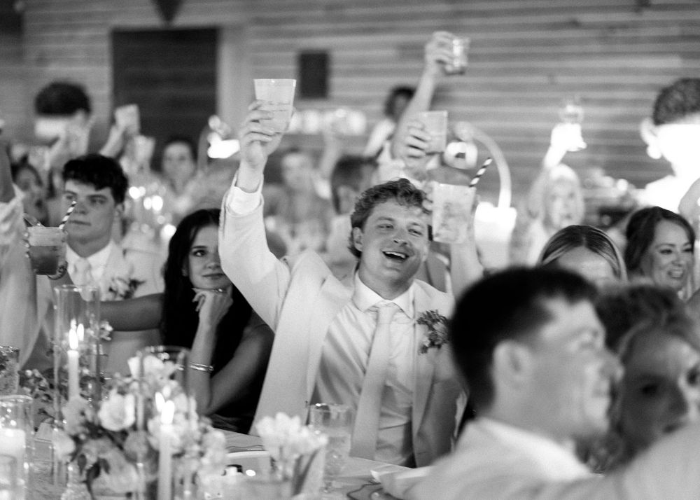 Joyful wedding guests raising glasses in a toast during reception celebration in rustic barn venue.