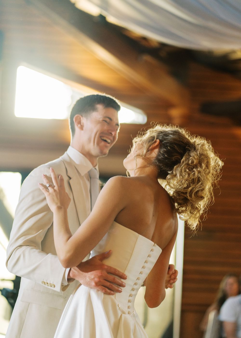 Bride and groom sharing first dance, laughing together in warm golden light at wedding reception.