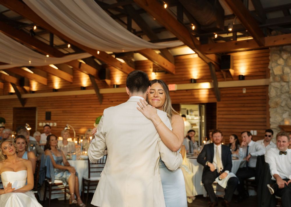 Bride and groom share their first dance in a rustic wood-beamed reception hall with guests watching.