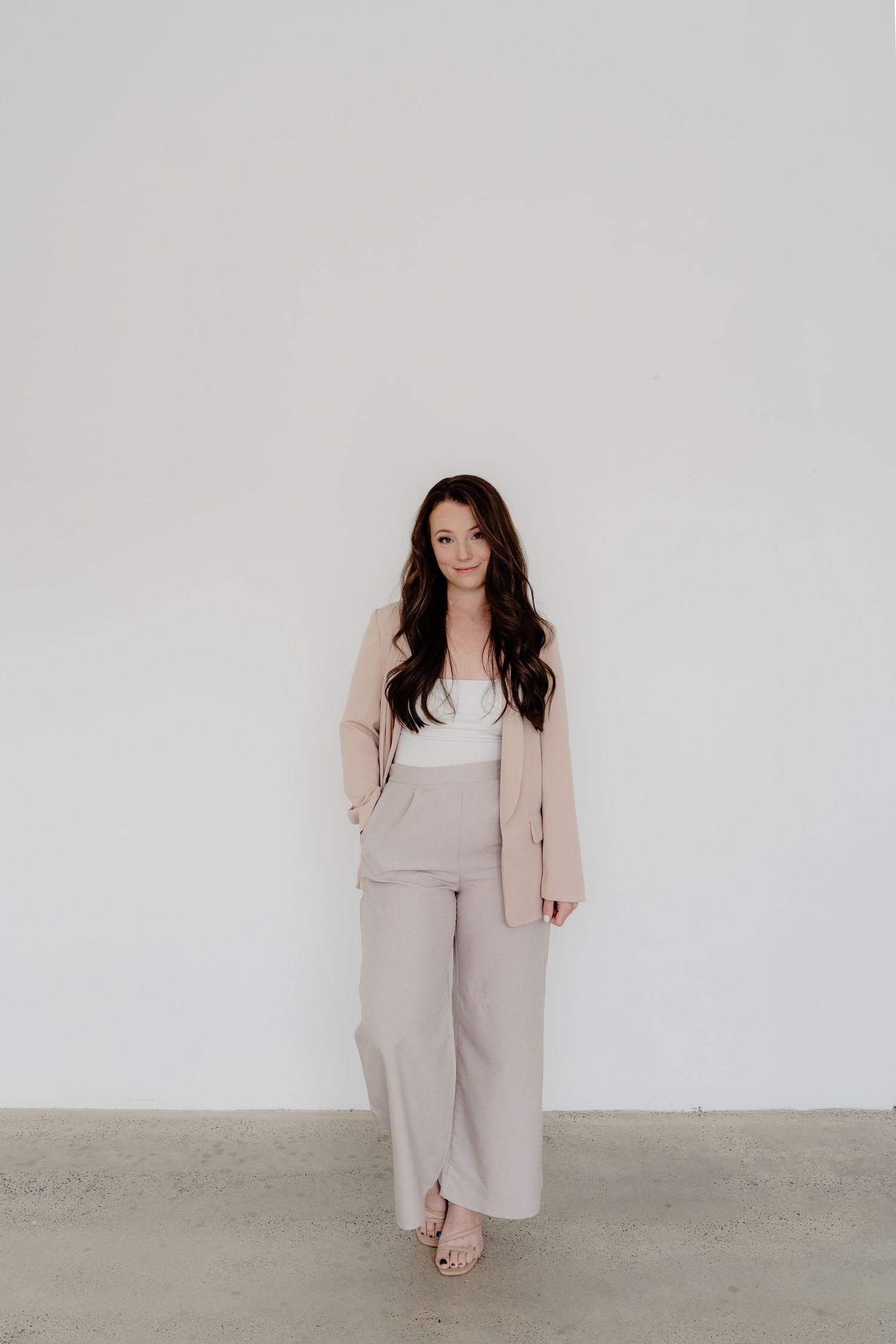 Woman in beige business suit and white top stands against plain white wall.