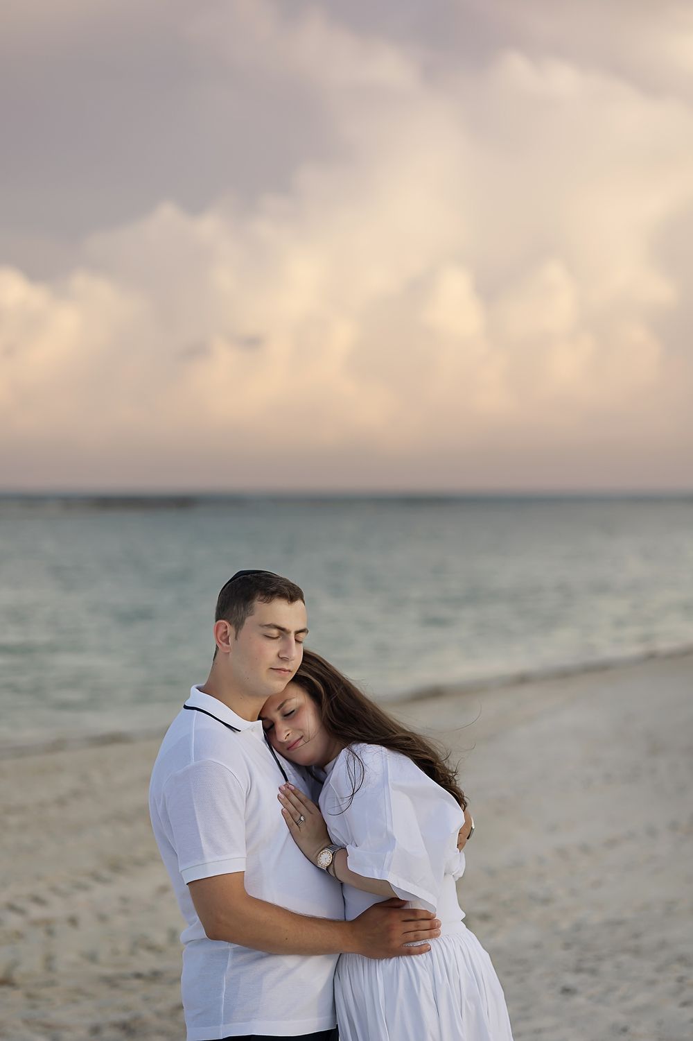 A romantic couple dressed in white embraces on a serene beach at sunset.